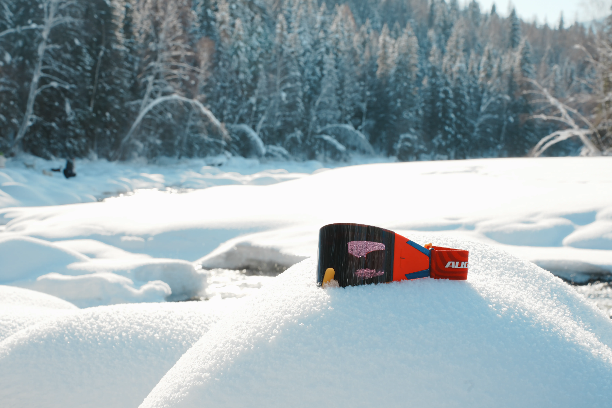 Red AUGMAX goggles with a red stripe and white graphic details resting on a snowy mound in a winter forest setting.